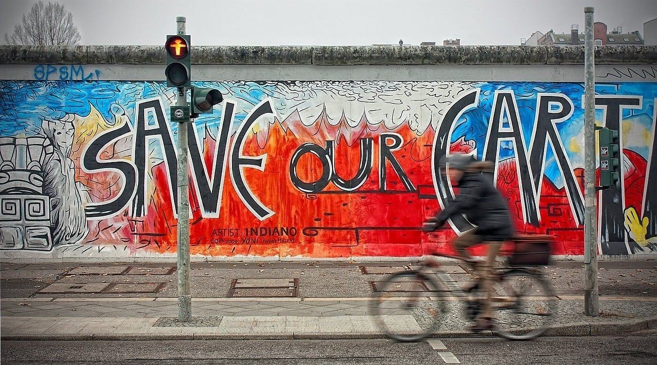 raffiti-Kunstwerk auf einer Mauer mit der Botschaft 'Save Our Art' in großen Buchstaben, farbenfroher Hintergrund mit Feuer- und Wolkeneffekten, während ein Fahrradfahrer vorbeifährt.