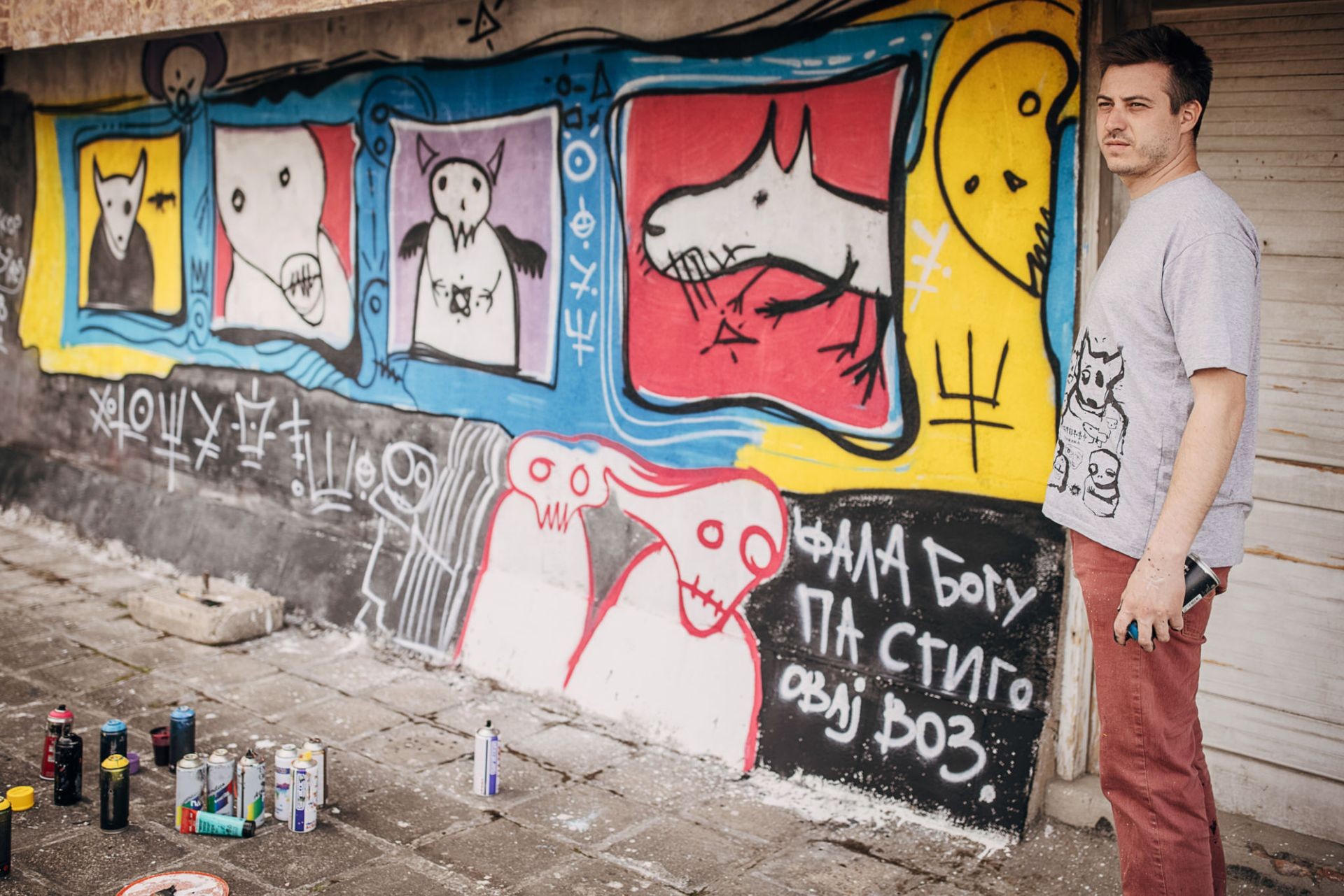 Straßenkünstler steht vor seiner bunten Graffiti-Wand mit abstrakten und surrealen Figuren, während Spraydosen auf dem Boden verteilt sind.